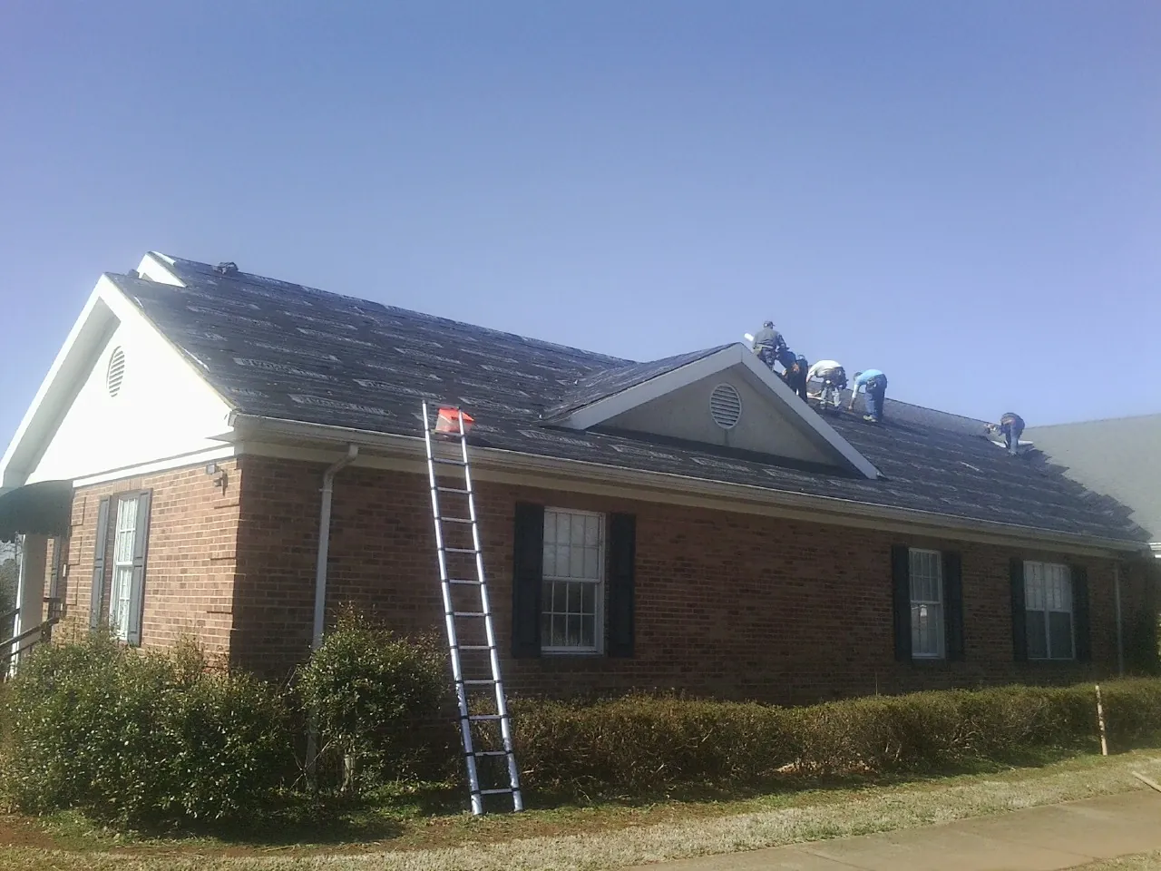 Crew tearing shingles off a brick ranch roof with a ladder set — Emergency Roof Repair work by our crew in Pike Road