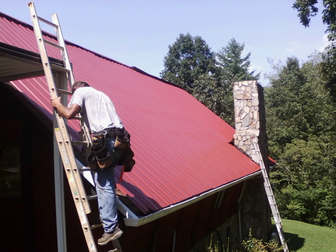 Roofer on a ladder beside a red metal roof and stone chimney — Soffit & Fascia Repair by Roofing Brothers in Pike Road